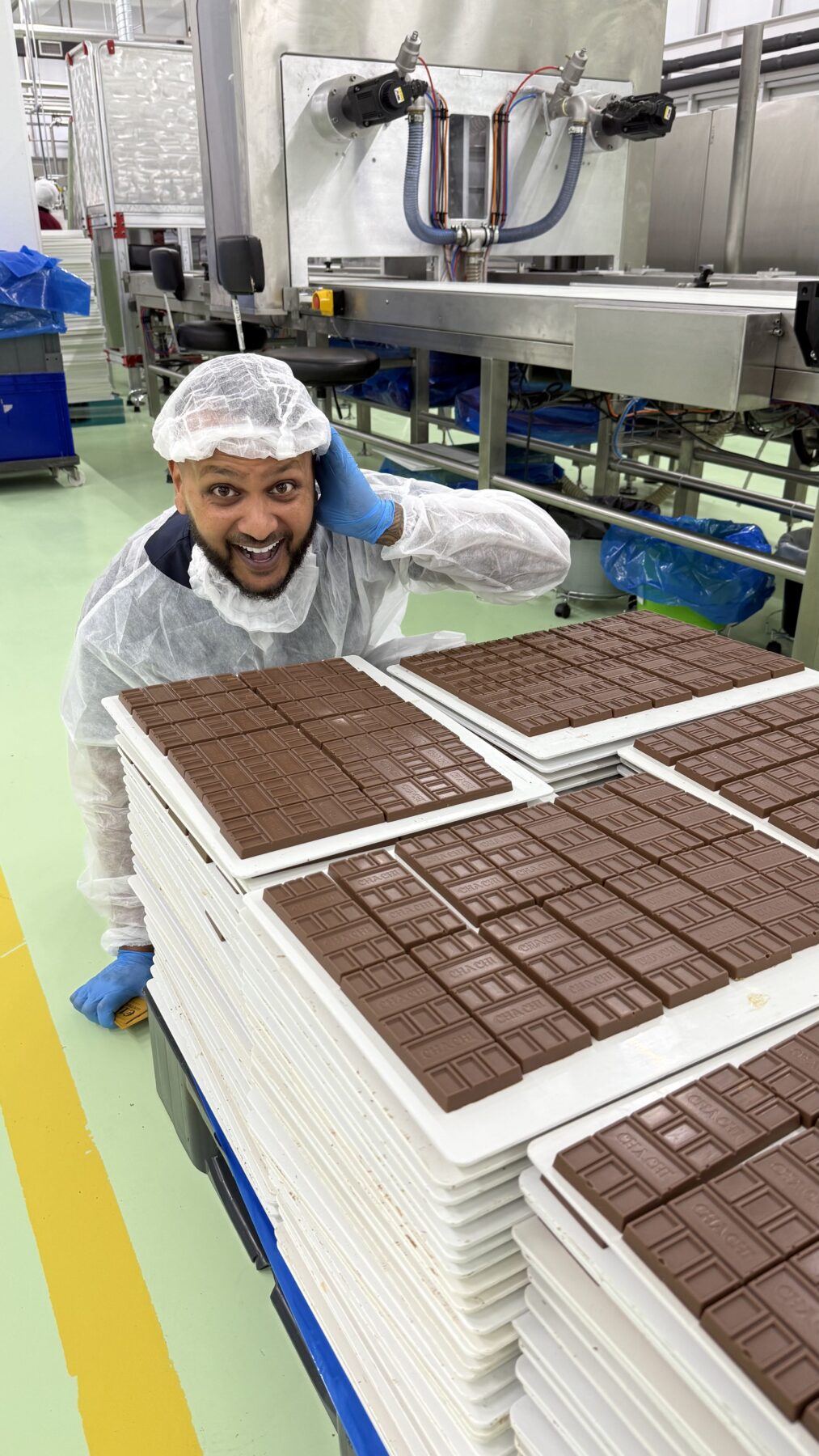 Cha Chi Versaci naast stapels platen met verse CHACHI-repen, het logo zichtbaar in de chocolade.